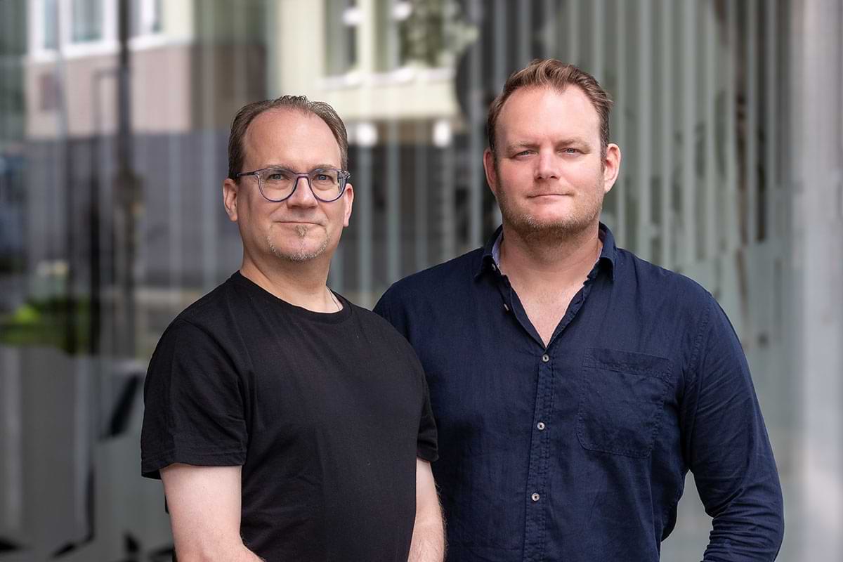 Zwei Männer: Markus Rykalski, links, trägt ein schwarzes T-Shirt und eine Brille. Stephan Raithel, rechts, trägt ein dunkelblaues Hemd. Beide schauen in die Kamera. Der Hintergrund ist unscharf.